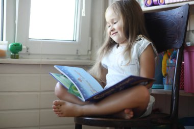 Küçük kız odada oturuyor ve kitap okuyor.
