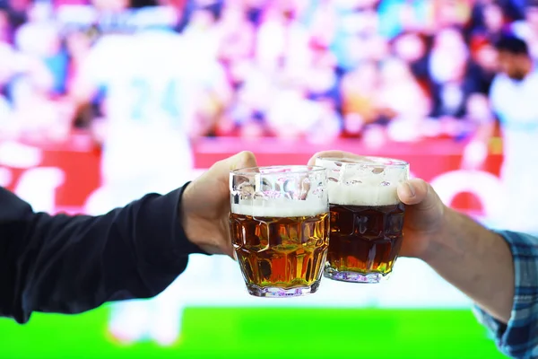 Hands horizontal holding lager beer glass and clinking on background of football game. Sport fans cheer up. Friends leisure lifestyle concept.