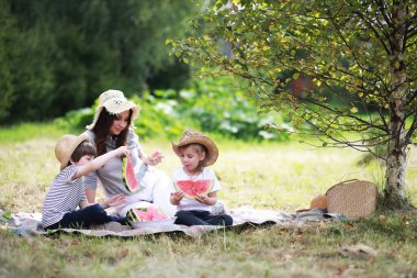 Parkta piknik yapan mutlu bir aile, bahçedeki çimlerde oturan ve dışarıda karpuz yiyen çocukları olan bir aile.