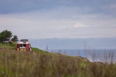 Vanlife ya da van life dolu dolu. Bulutlu bir günde, doğanın ortasına park etmiş, arkasında görünür yatağı olan eski kırmızı bir minibüs. Minibüsle özgürlük.