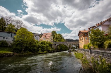 Mura ya da Mur nehri bulutlu ama güneşli bir günde Avustralya 'nın orta kesiminde Murau şehir merkezinden akar. Arka planda taş köprü ve klasik binalar görülüyor.