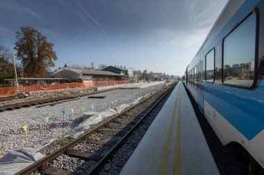 Train station is being renovated, trains stopped at a station and a closed train track with freshly built rails and sleepers is visible. Work train on tracks.