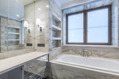 The original design of the bathroom with beige marble tiles.A large mirror and a bathtub by the window.