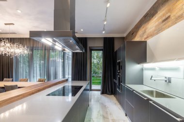 The interior of a stylish gray kitchen with light countertops, a hood over the hob. Nice view on a backyard.