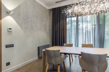 The interior of a stylish living room with armchairs around a large table and a luxurious chandelier against a gray wall and window.