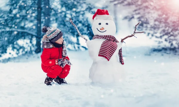 Gyerek ül a hóban, közel egy vicces hóember — Stock Fotó