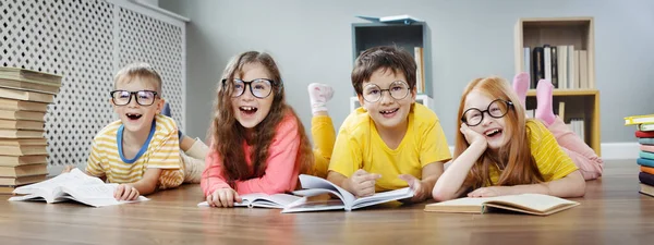 Quatro crianças bonitos em óculos deitados no chão dentro de casa com livros — Fotografia de Stock