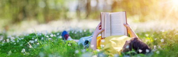 Маленький ребенок лежит на поле в весеннем парке и читает книгу — стоковое фото
