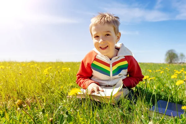 Little child sitting with book on the field with dandelions. — Fotografia de Stock