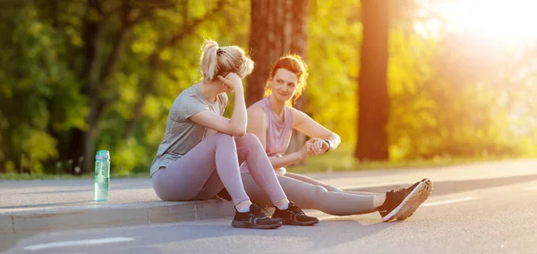 Two women sitting on the border of the roadside after a run. Panoramic view. — Stockfoto