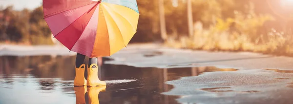 Child standing in the puddle in yellow rubber boots and holding colourful umbrella in hands. Concept of childhood.