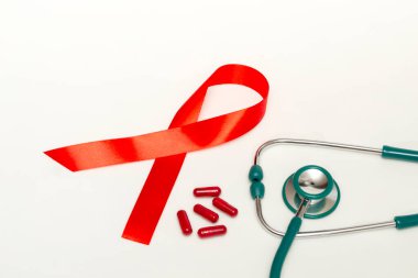 Dünya AIDS Günü Aralık ayı için tıbbi konsept. Kırmızı kurdele, yeşil tıbbi steteskop ve beyaz arka planda kırmızı terapi hapları veya vitaminler. Kopyalama alanını kapat
