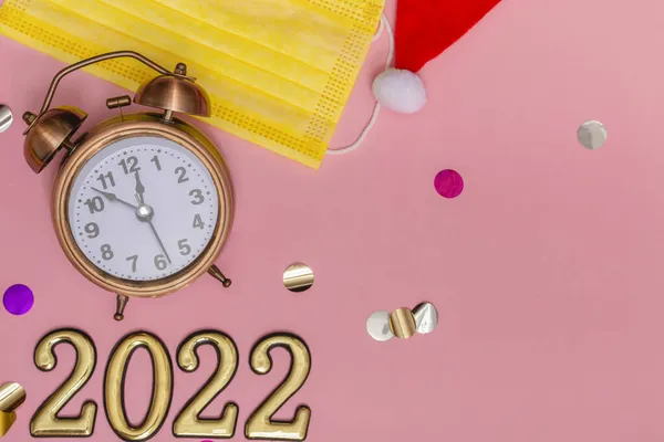 Yeni yıl konsepti pembe arka plan alarm saati sarı tıbbi maskeli, altın numaralar 2022, kapatın.
