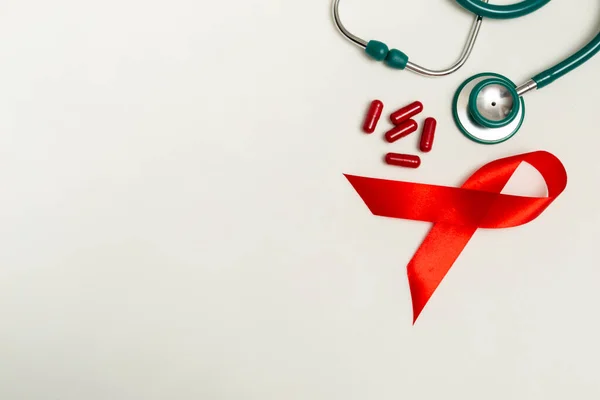 Dünya AIDS Günü Aralık ayı için tıbbi konsept. Kırmızı kurdele, yeşil tıbbi steteskop ve beyaz arka planda kırmızı terapi hapları veya vitaminler. Kapat.