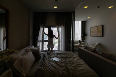 Güzel bir kadın perdeleri açıyor ve pencereden bakıyor.