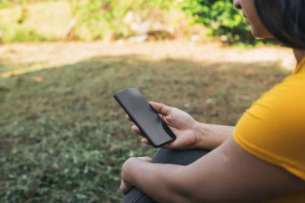 Cep telefonuyla sahada oturan Latin bir kadın, cep telefonunu doğa ortamında tutan bir kadın.