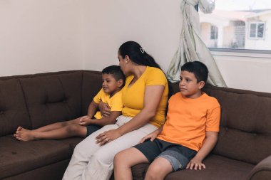 Anne oğluna sarılıyor ve onu öpüyor, koltukta oturuyor.