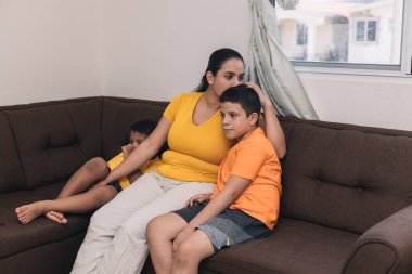 Anne oğluna sarılıyor ve onu öpüyor, koltukta oturuyor.