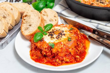 Yumurtalı Shakshuka, İtalyan sosisi, domates sosu, peynirli ve fesleğenli, beyaz bir tabakta, yatay