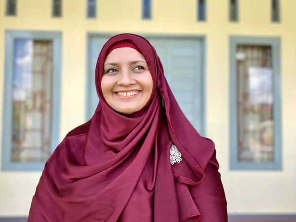 Asian woman wearing red hijab looking away smiling in front of house