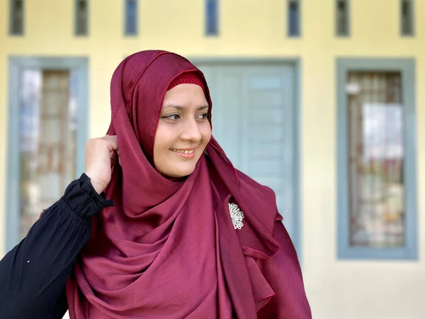 Asian woman wearing red hijab looking away smiling in front of house