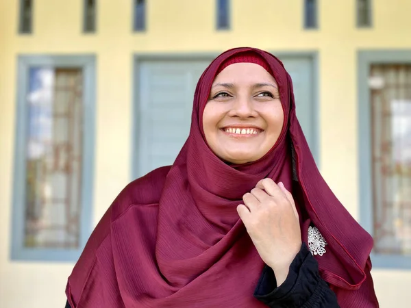 Asian woman wearing red hijab looking away smiling in front of house