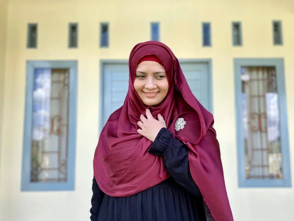 Asian woman wearing red hijab looking away smiling in front of house