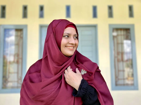 Asian woman wearing red hijab looking away smiling in front of house
