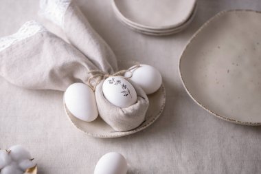 Beyaz paskalya yumurtaları bej plakalı, siyah filizli. Masanın üzerinde keten bir masa örtüsü ve bir pamuk peçete var. Doğal malzemeler, minimum konsept. Metin için yer.