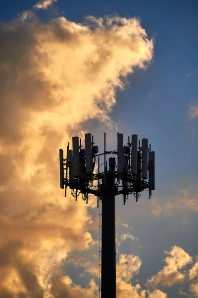 The upper part of a cellular tower is seen as the setting sun lights up a storm cloud rolling in behind it.