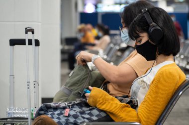 Bangkok, Tayland - 16 Ocak 2022: Koronavirüs veya covid virüs için koruyucu yüz maskesi takan Asyalı genç kız havaalanı terminalinde uçuş beklerken akıllı telefondan müzik dinliyor..