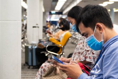 Bangkok, Tayland - 16 Ocak 2022: Coronavirus veya covid virüsü için koruyucu yüz maskesi takan Asyalı genç yolcu ve havaalanı terminalinde beklerken havayolu uygulamasında uçuş gecikmesini kontrol etti.