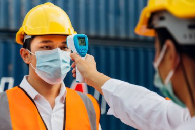 Surat maskesi takan Asyalı işçi inşaat alanındaki covid, covid-19 veya coronavirus salgınından korunmak için işe girmeden önce ateşi dijital termometreyle kontrol ediyor. seçici odak.