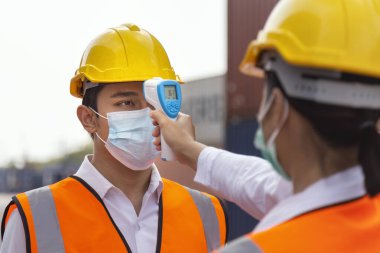 Surat maskesi takan Asyalı işçi inşaat alanındaki covid, covid-19 veya coronavirus salgınından korunmak için işe girmeden önce ateşi dijital termometreyle kontrol ediyor. seçici odak.