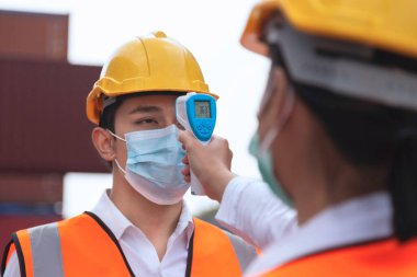 Surat maskesi takan Asyalı işçi inşaat alanındaki covid, covid-19 veya coronavirus salgınından korunmak için işe girmeden önce ateşi dijital termometreyle kontrol ediyor. seçici odak.