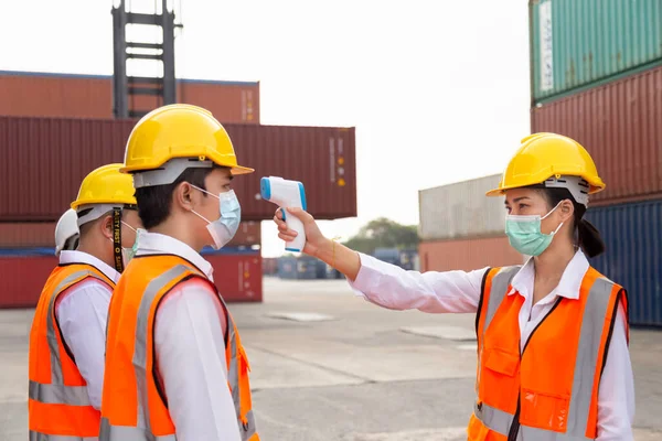 İnşaat alanındaki kovid, covid-19 veya koronavirüs salgınından korunmak için işe girmeden önce yüz maskesi takan Asyalı işçiler ateşi dijital termometreyle kontrol ediyorlar. seçici odak.