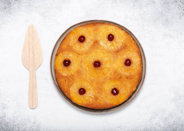 Homemade pineapple upside down cake with glace cherries and  wooden pie server on light rustic background. Top view. Copy space.