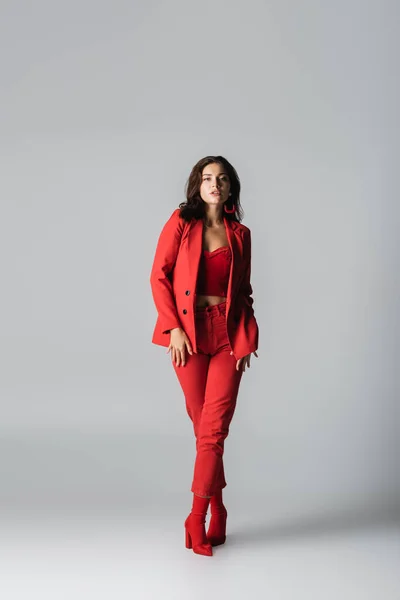 full length of young trendy woman in red boots and suit posing on grey