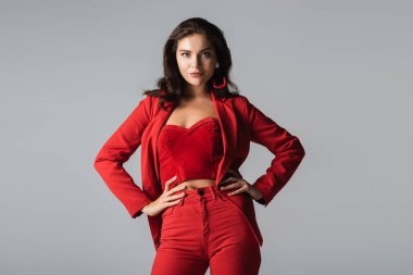 Kırmızı takım elbiseli, küpeli, elleri kalçasında poz veren gri elbiseli genç bir kadın.