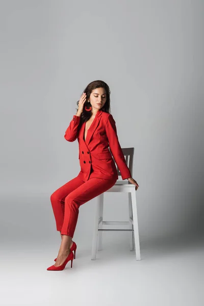 full length of trendy woman in red suit posing while sitting on white chair on grey