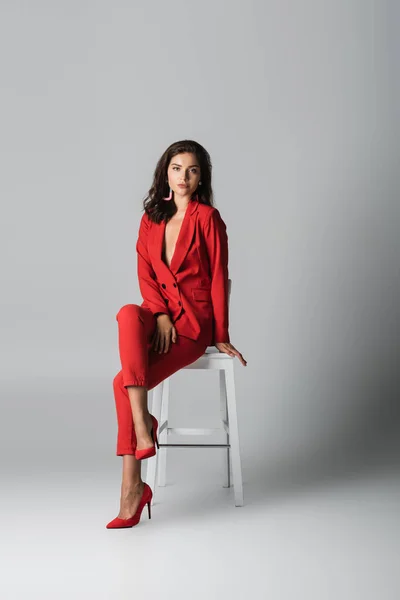 full length of trendy woman in red stylish suit posing while sitting on white chair on grey