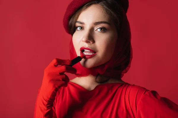 スカーフ ベレー帽の若い女性は赤で隔離された口紅を適用します — ストック写真