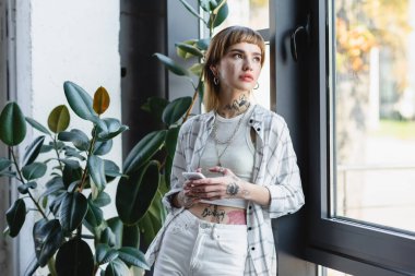 Ofisin penceresinin yanında duran akıllı telefonu tutan dövmeli genç bir kadın.