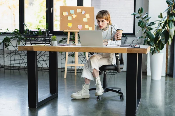 stylish tattooed businesswoman holding coffee to go while sitting at desk near laptop