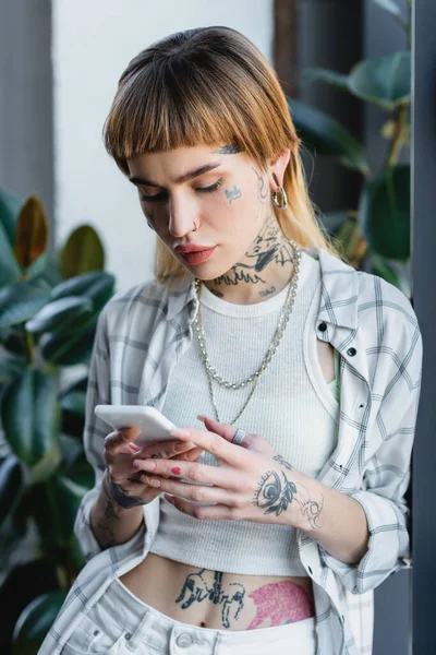 tattooed woman in silver necklaces and piercing messaging on mobile phone in office