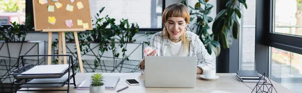 smiling businesswoman with tattoo pointing with hand during video call on computer, banner