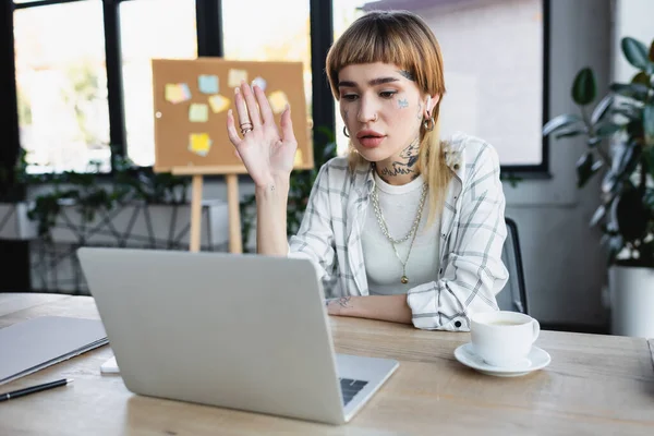 trendy tattooed businesswoman waving hand during video call on laptop in office