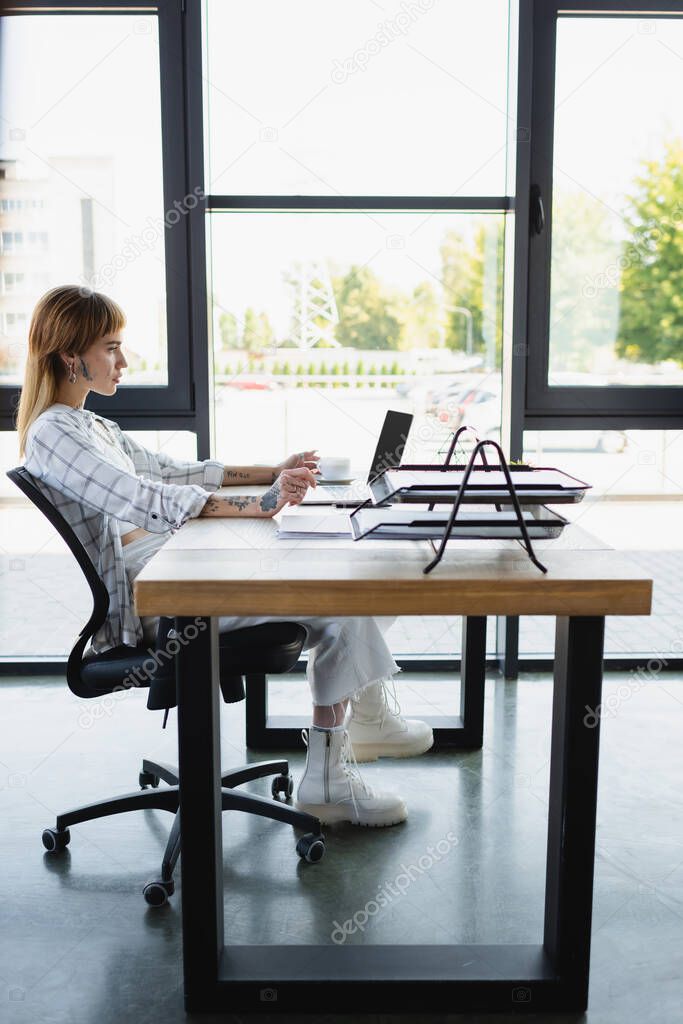 side view of young and stylish woman sitting at work desk near laptop with blank screen