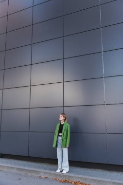 Açık havada yüksek gri duvarın yanında, dövmeli, modaya uygun kıyafetler giyen bir kadın.