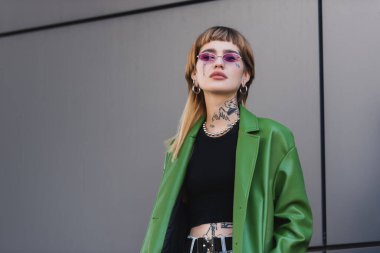 Gri duvarın yanında duran zarif elbiseli, dövmeli genç bir kadının alçak açılı görüntüsü.